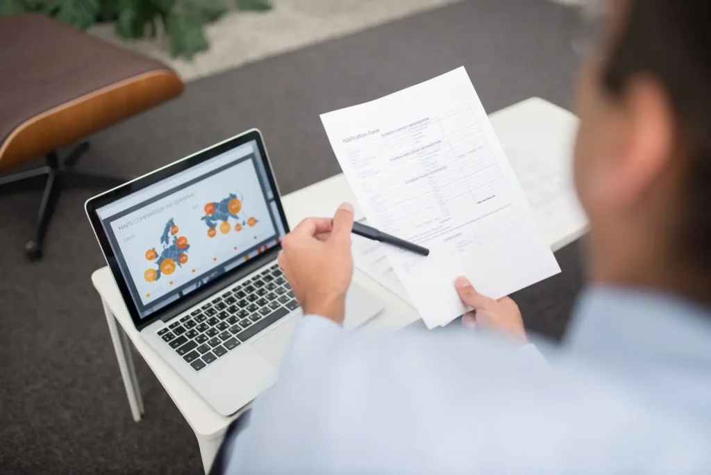 Professional reviewing documents next to a laptop showing charts